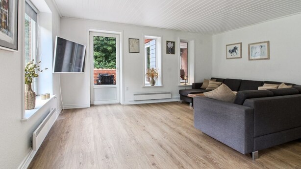 A spacious living room with a grey sofa, wooden flooring, and large windows offering views of the outdoors.