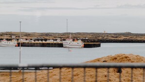 Cena costeira com barcos atracados no cais, um farol ao longe e uma praia arenosa em primeiro plano.