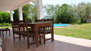 A covered patio with a wooden table and chairs, a small pool, and a grassy area with trees.