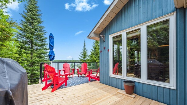 A wooden deck with red Adirondack chairs, a grill, and a large window overlooking a forested area.