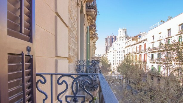 Un balcón con barandillas de metal decorativo que da a una calle con edificios y árboles.