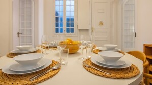 Una mesa de comedor dispuesta con platos, cuencos y vasos blancos, con un mantel tejido y un bol de fruta en el centro.