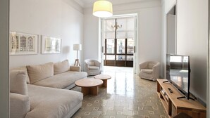 Un salón moderno con un sofá beige, dos sillones, una mesa de centro de madera, un televisor sobre un mueble bajo y un ventanal con vistas a edificios.