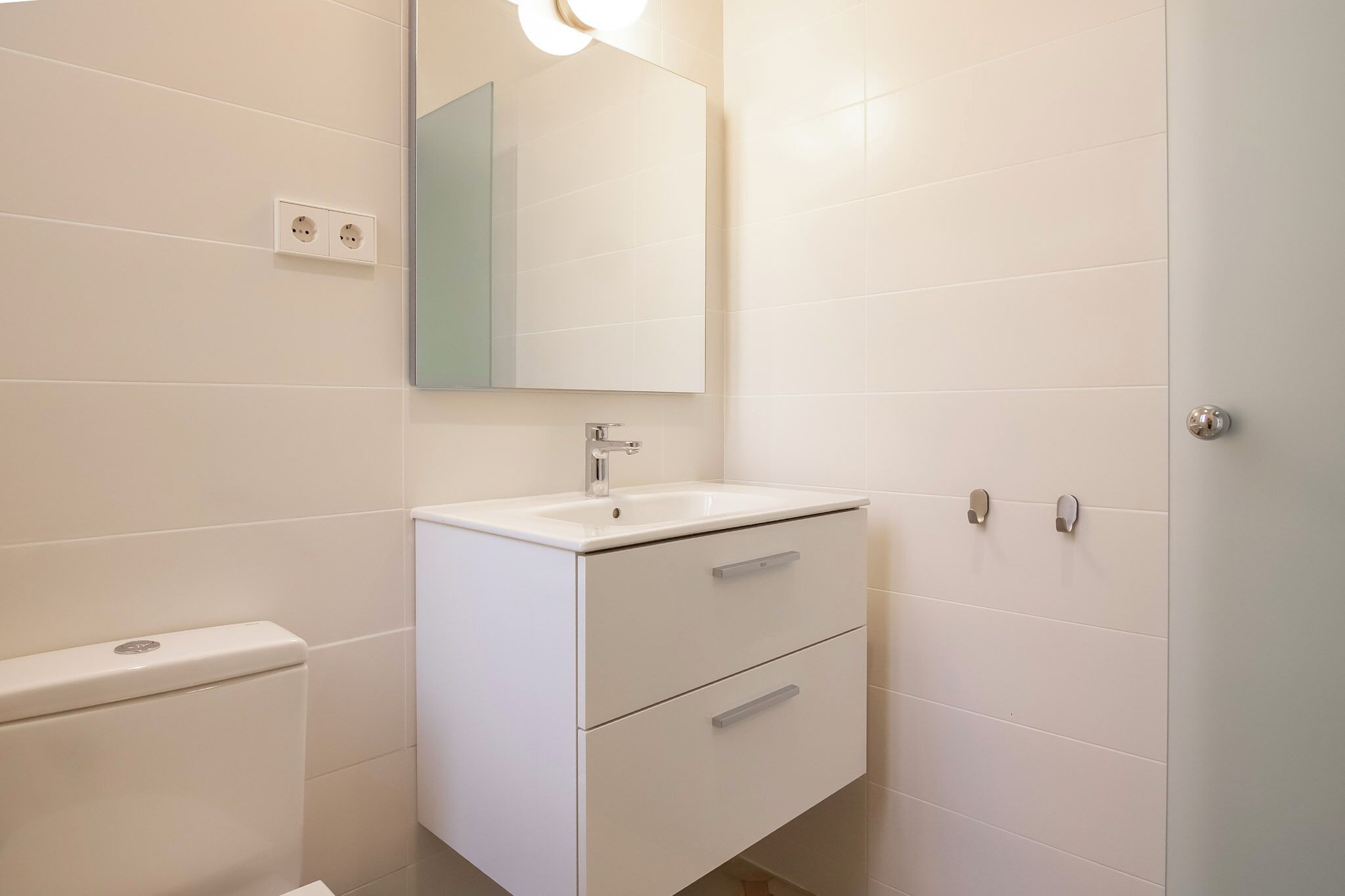 Ein Badezimmer mit einem weißen Waschbecken, einem Spiegel und zwei Schubladen. Eine Toilette befindet sich links, eine Tür rechts.