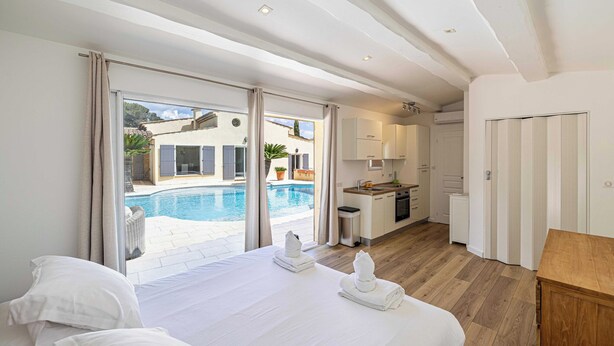 Ein Schlafzimmer mit einem großen Bett, Blick auf einen Swimmingpool und einer kleinen Küchenzeile im Hintergrund.