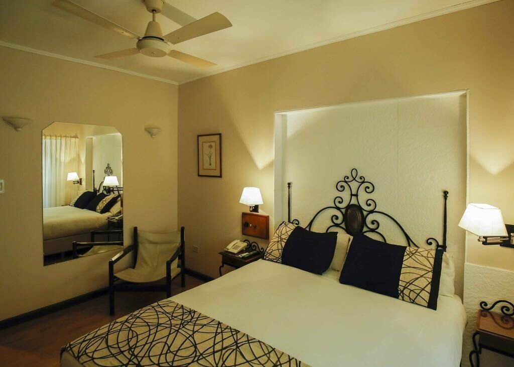 A hotel room with a large bed, a chair, a desk with a lamp, a telephone, and a mirror.