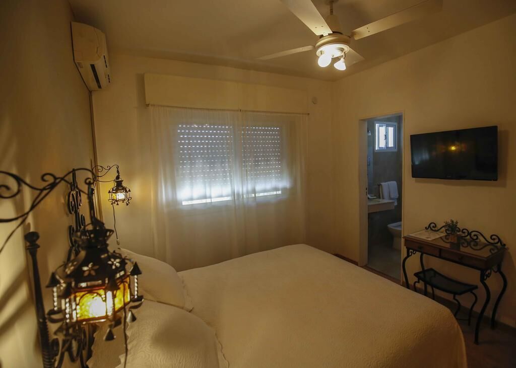 A bedroom with a bed, a ceiling fan, a window with blinds, a wall-mounted TV, and a small table with a plant.