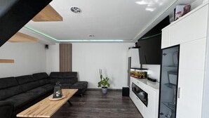 A modern living room with a sectional sofa, a wooden coffee table, a potted plant, and a kitchen area with white cabinets and a glass shelf. - Spacious Retreat for 12 With Spa Option (Saint-Mars-d'Outille)