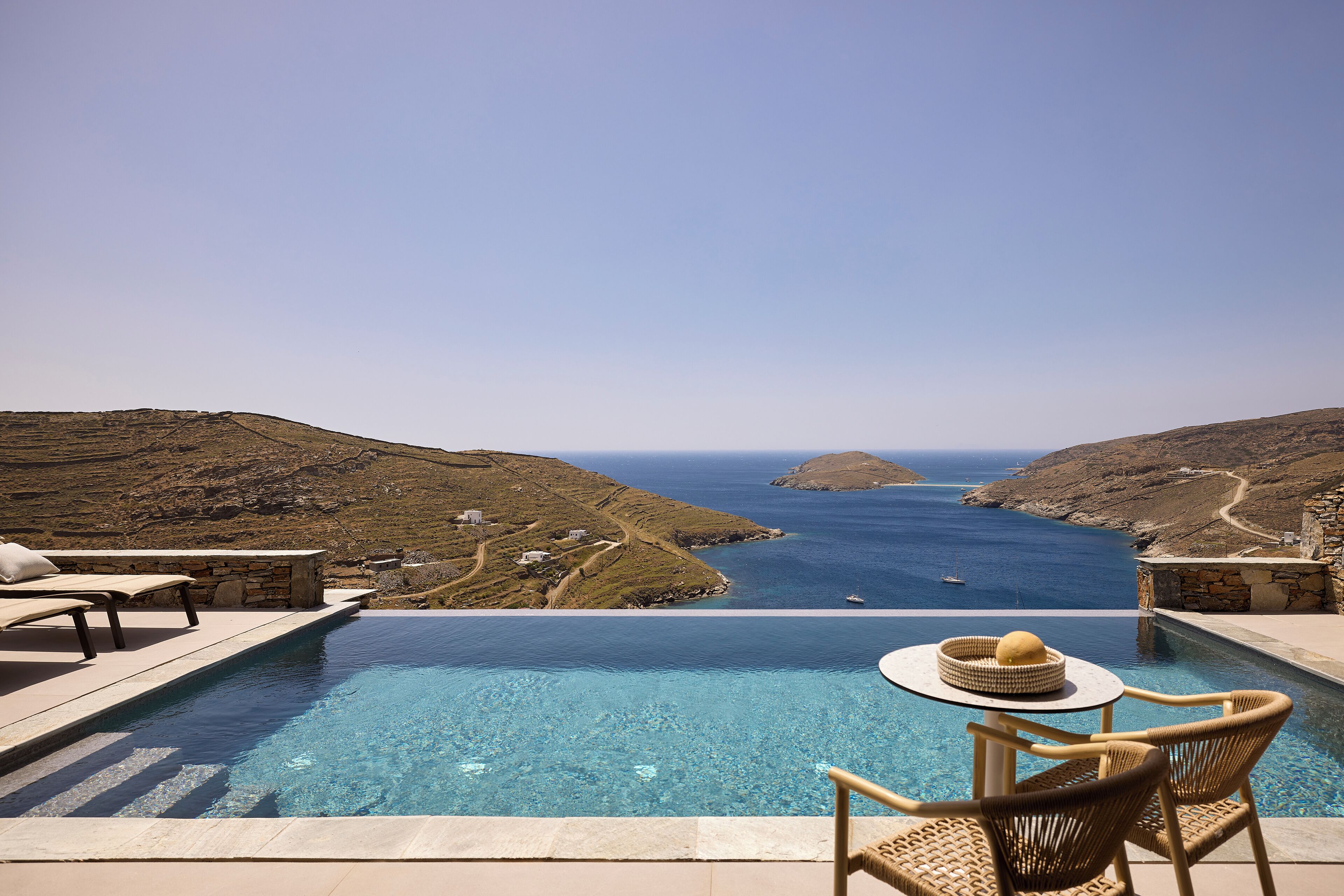 Una piscina infinita con vistas al mar y colinas, mobiliario exterior y una estructura de piedra.