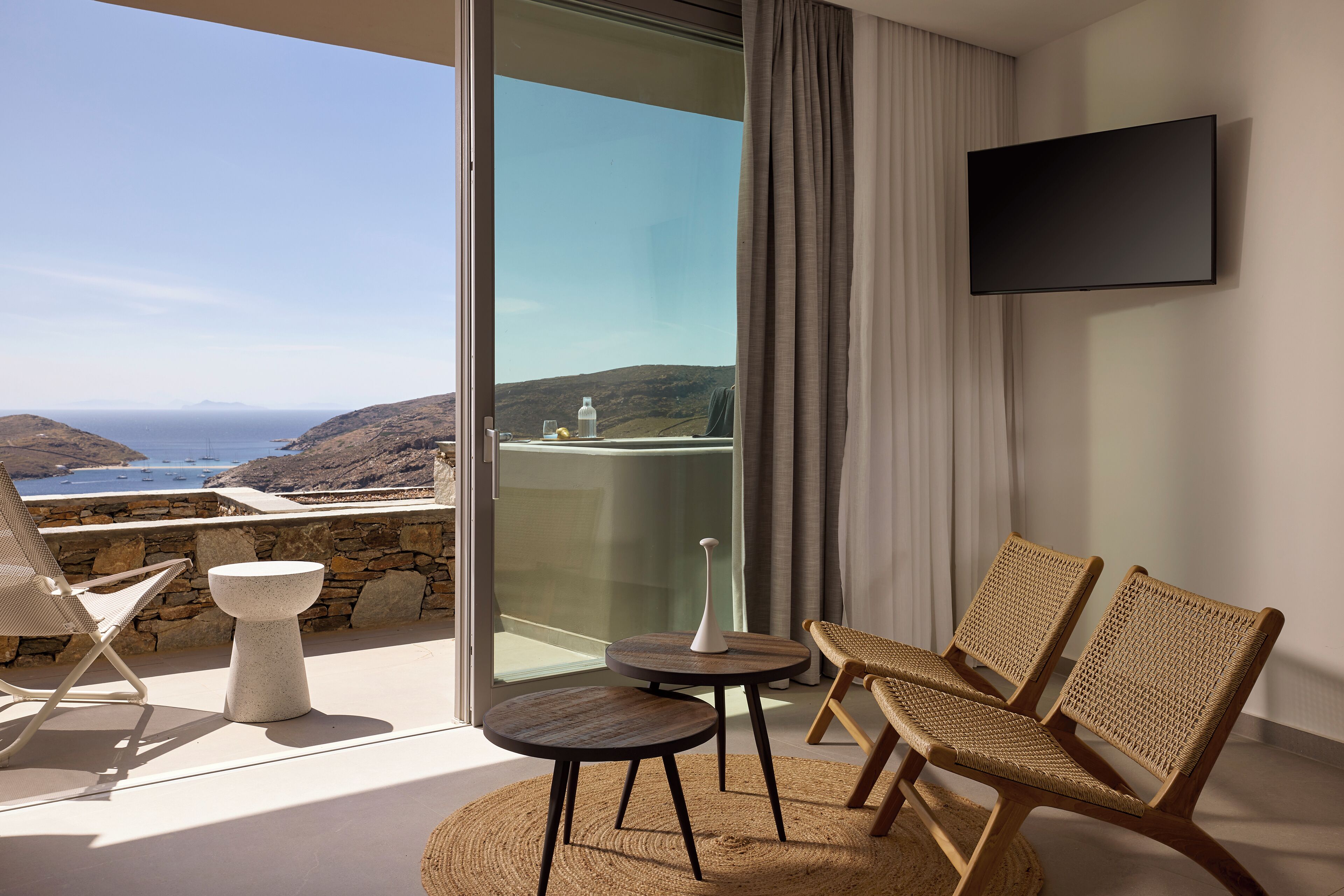 Un balcón con vista al mar, dos sillones de mimbre, una mesita y un televisor de pantalla plana montado en la pared.