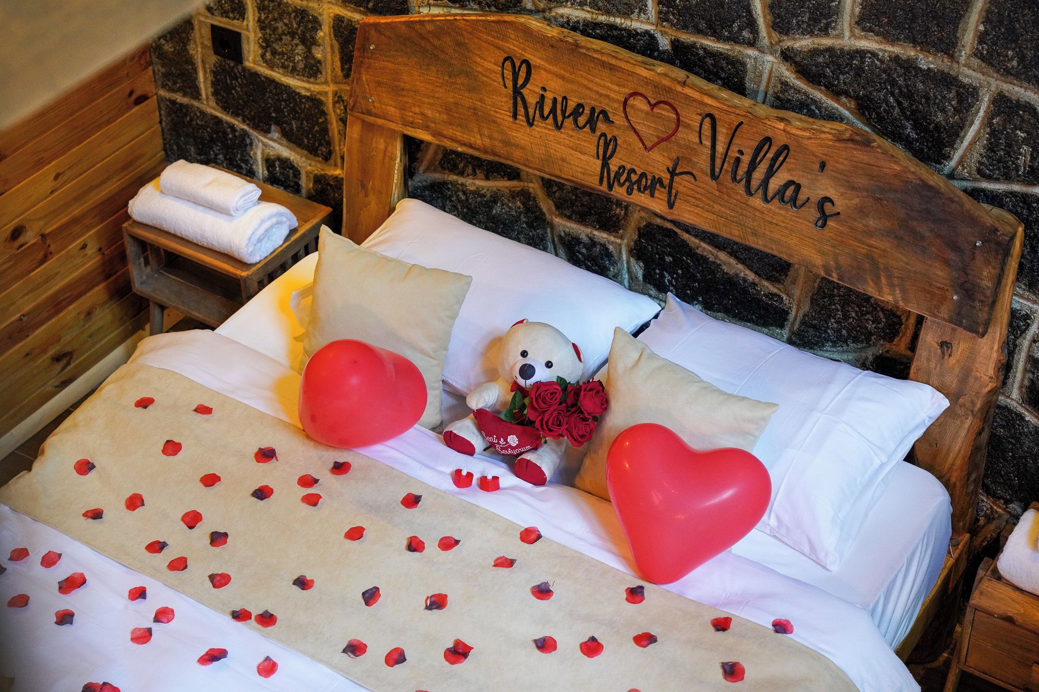 Una cama con cabecera de madera, ropa de cama blanca, un oso de peluche, rosas rojas y globos con forma de corazón.