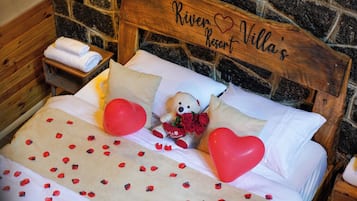 Una cama con cabecera de madera, ropa de cama blanca, un oso de peluche, rosas rojas y globos con forma de corazón.