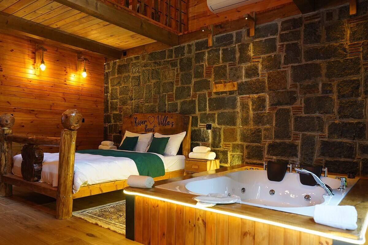 A wooden cabin-style bedroom with a stone wall, a bed with a headboard reading "River Heart Retreat," and a large bathtub with a wooden bench.