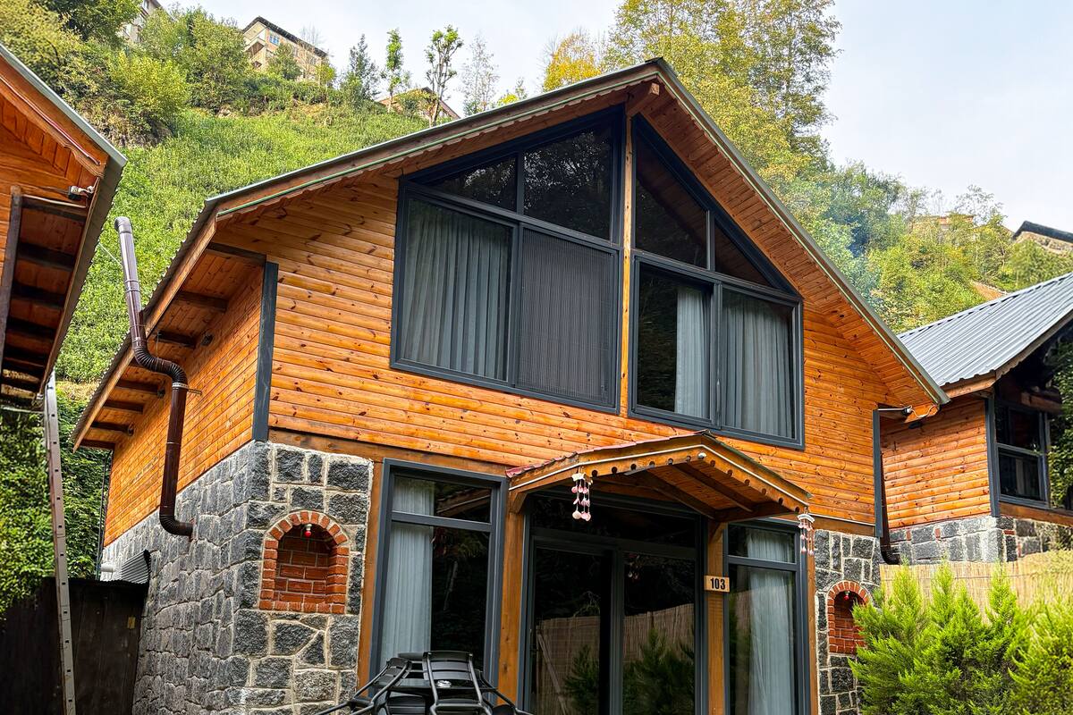 A two-story wooden house with a stone foundation, large windows, and a number '103' on the front door.