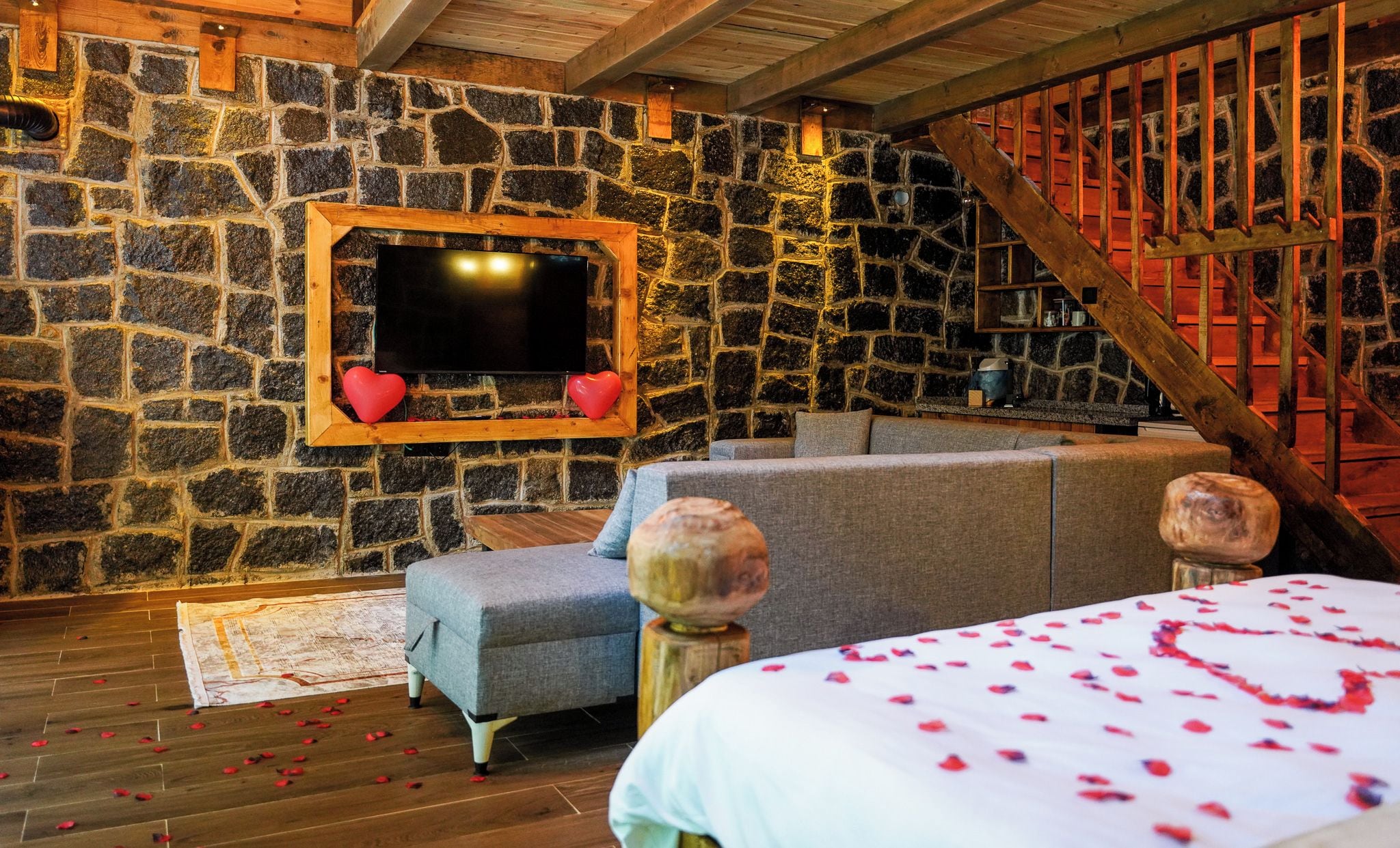 Quarto aconchegante com parede de pedra, televisão de tela plana, sofá e cama com pétalas de rosa.