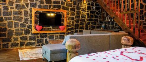 Quarto aconchegante com parede de pedra, televisão de tela plana, sofá e cama com pétalas de rosa.