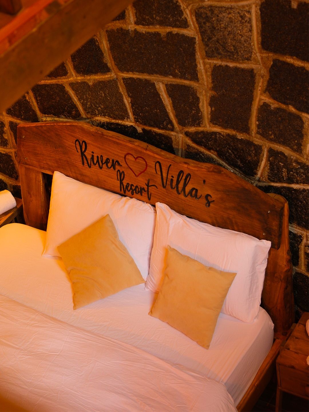 A bed with a headboard labeled 'River Resort Villa's' against a stone wall backdrop.