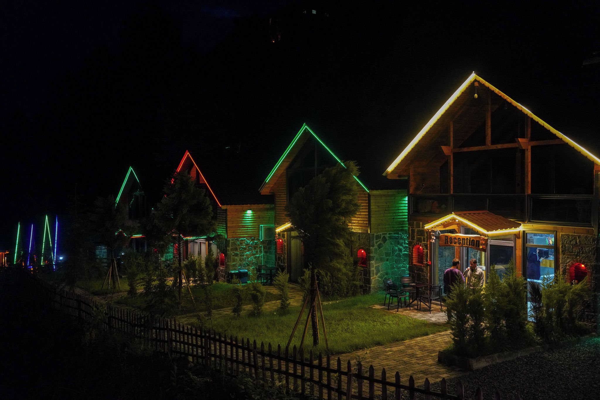 A row of houses with illuminated roofs, a wooden fence, and a small outdoor seating area.