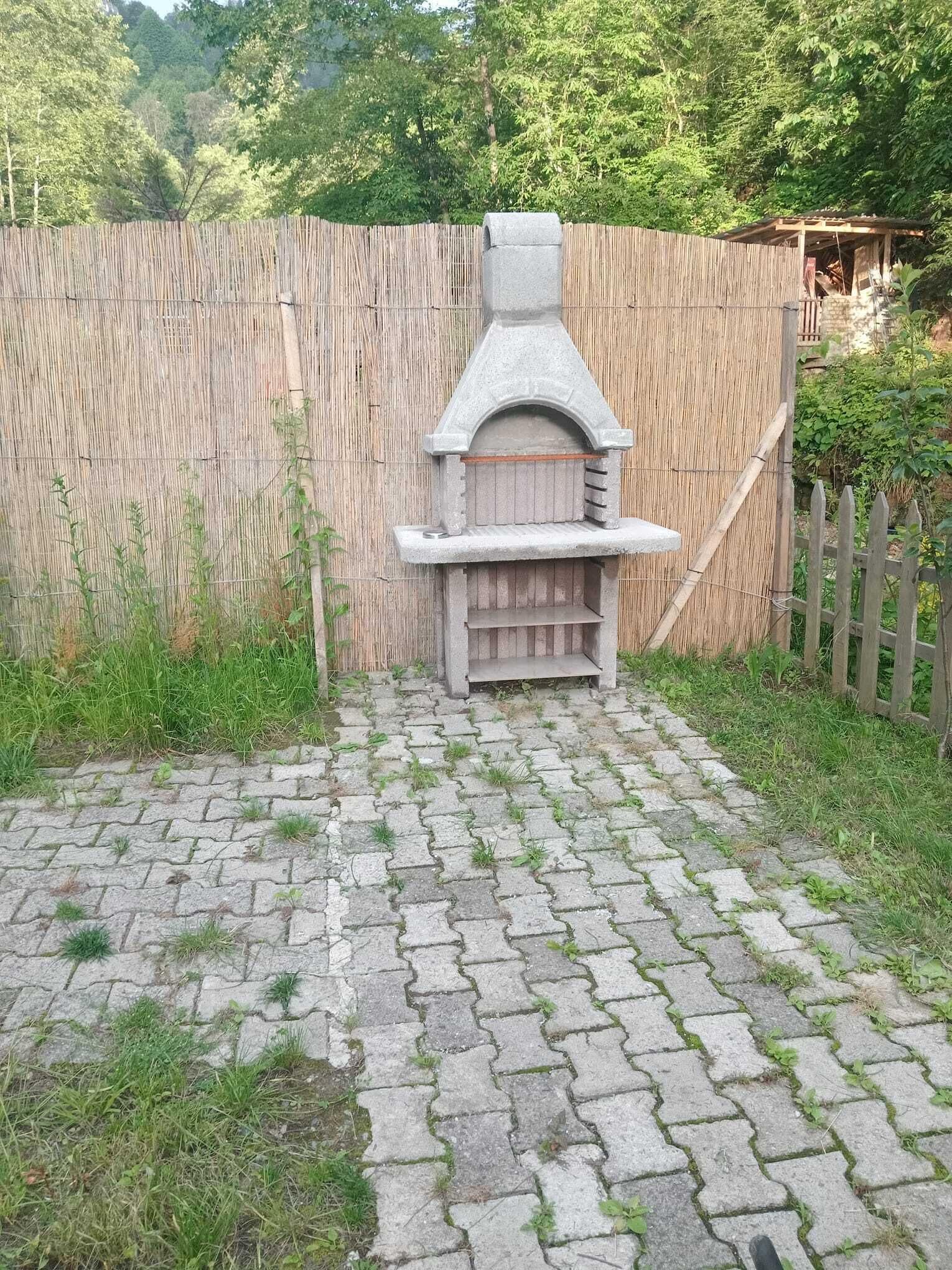 A stone outdoor grill with a chimney, surrounded by a bamboo fence and a wooden fence, set on a paved area with grass patches.
