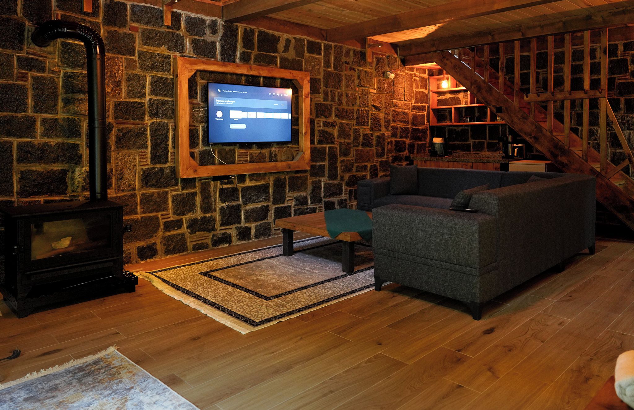 A cozy living room with a stone wall, a fireplace, a television mounted on the wall, a sofa, and a staircase.