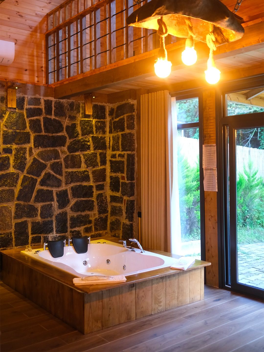 A wooden cabin interior with a stone wall, a large bathtub, and a wooden ceiling with hanging lights.