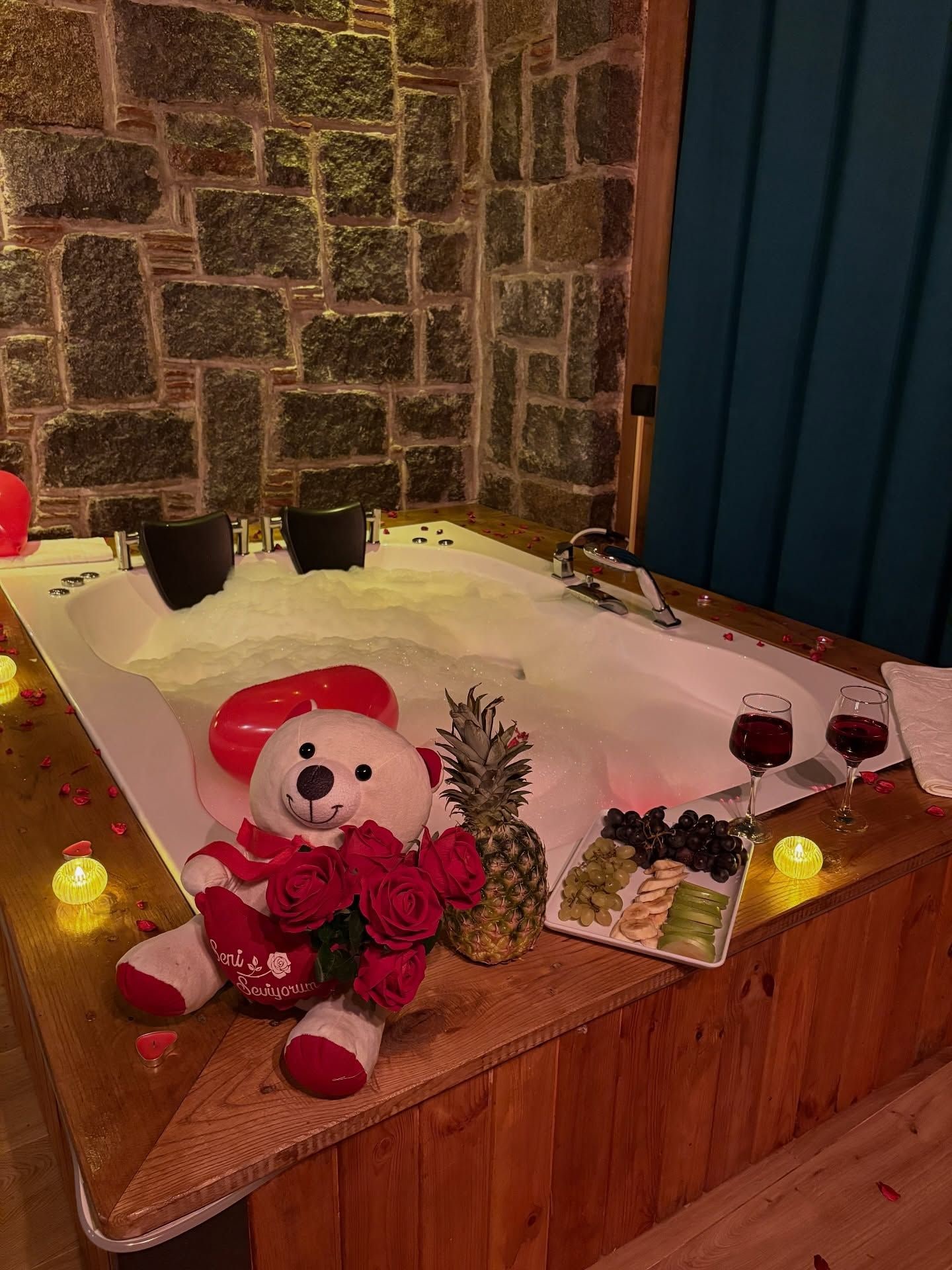 A wooden bathtub with a stone wall backdrop, a plush teddy bear holding roses, a pineapple, and a fruit platter with grapes and bananas.