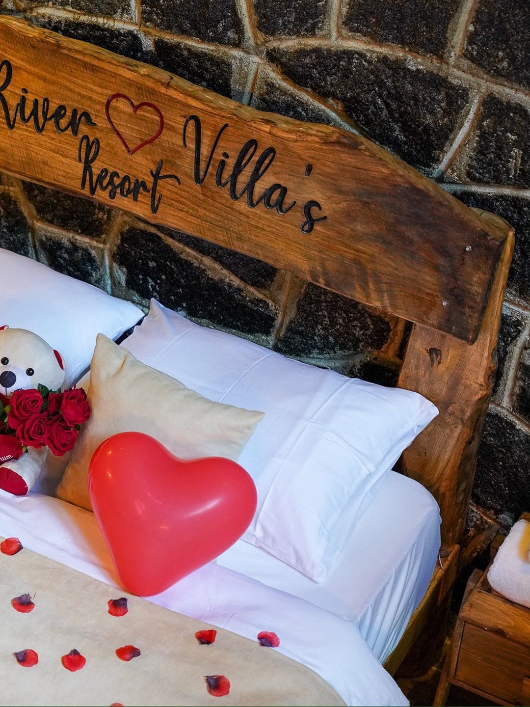 Ein Bett mit Holz-Kopfteil, weißer Bettwäsche, einem Teddybären, roten Rosen und einem herzförmigen Kopfkissen in Rot.