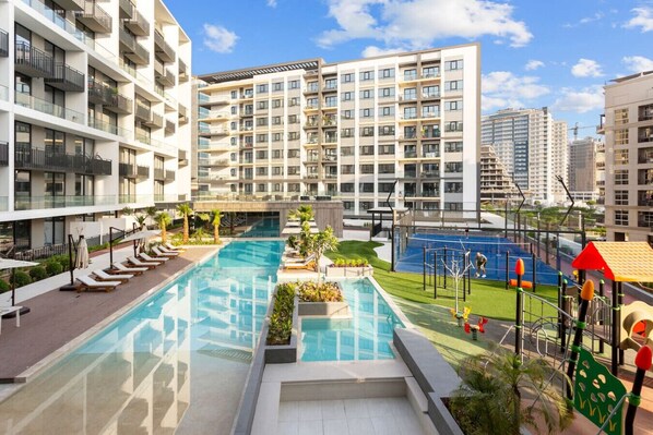 A modern apartment complex with a central swimming pool, outdoor seating, and a playground.