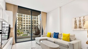Un salon moderne comprenant un canapé, un fauteuil, un téléviseur à écran plat et une grande fenêtre donnant sur un immeuble d’habitation.