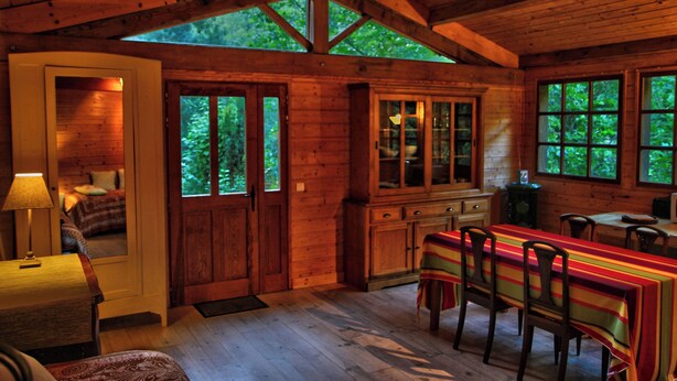 A wooden cabin interior with a dining area, kitchen, and a large window overlooking greenery. - Atelier du Moulin all in wood classified 2 * for 2 to 4 pers - on the banks of the torrent (Aubusson-d'Auvergne)