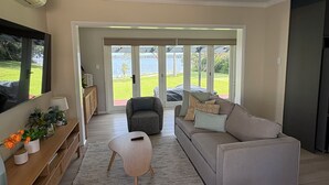 A spacious living room with a grey sofa, a wooden coffee table, and a grey armchair. There is a large window with a view of a green lawn and a body of water.