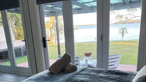 A bedroom with a large bed, a view of the outdoors through glass doors, and a patio area with a table and chairs.