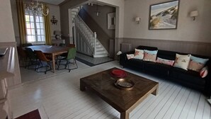 Een woonkamer met een houten salontafel, een bank met opvallende kussens, een eethoek met een tafel en stoelen, en een trap naar een hoger niveau.