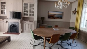 Een eetruimte met een ronde tafel, stoelen, een televisie, keukenkastjes, een open haard en een schilderij aan de muur.