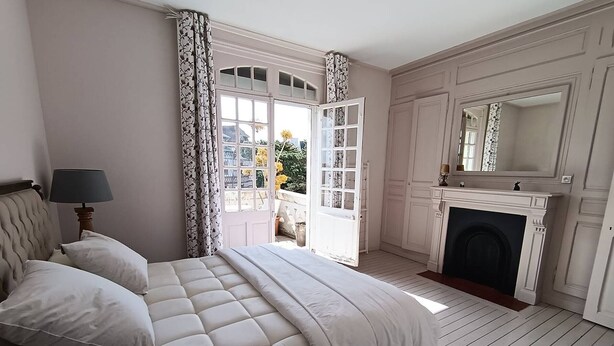 Een slaapkamer met een bed, nachtkastje, lamp, open haard en een groot raam met gordijnen.