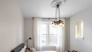 A bedroom with a large bed, a hanging light fixture, a window with curtains, and a small window with a stained glass design.