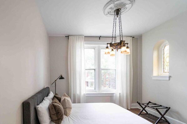 Une chambre avec un grand lit, un luminaire suspendu, une fenêtre avec des rideaux et une petite fenêtre ornée de vitraux.