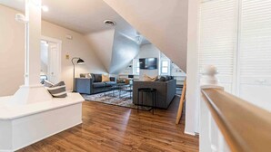 A modern loft apartment with a high ceiling, wooden flooring, a grey sofa set, a flat-screen TV, and a large window.