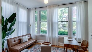 Ein Wohnzimmer mit einem braunen Ledersofa, einem Holztisch und einem großen Fenster mit Vorhängen.