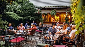 An outdoor seating area with round tables and chairs, surrounded by greenery and a building with warm lighting. - 1BR Condo Steps to the Monastery, Cafes, and More (Cincinnati)