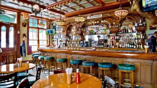 A bar with wooden bar stools, a counter with bottles, and a sign that reads "No Sniveling". - 1BR Condo Steps to the Monastery, Cafes, and More (Cincinnati)