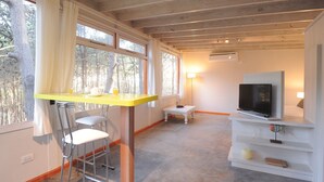 Une pièce avec un plafond en bois, une table jaune avec des chaises, un téléviseur posé sur un meuble blanc et une petite table avec une lampe.