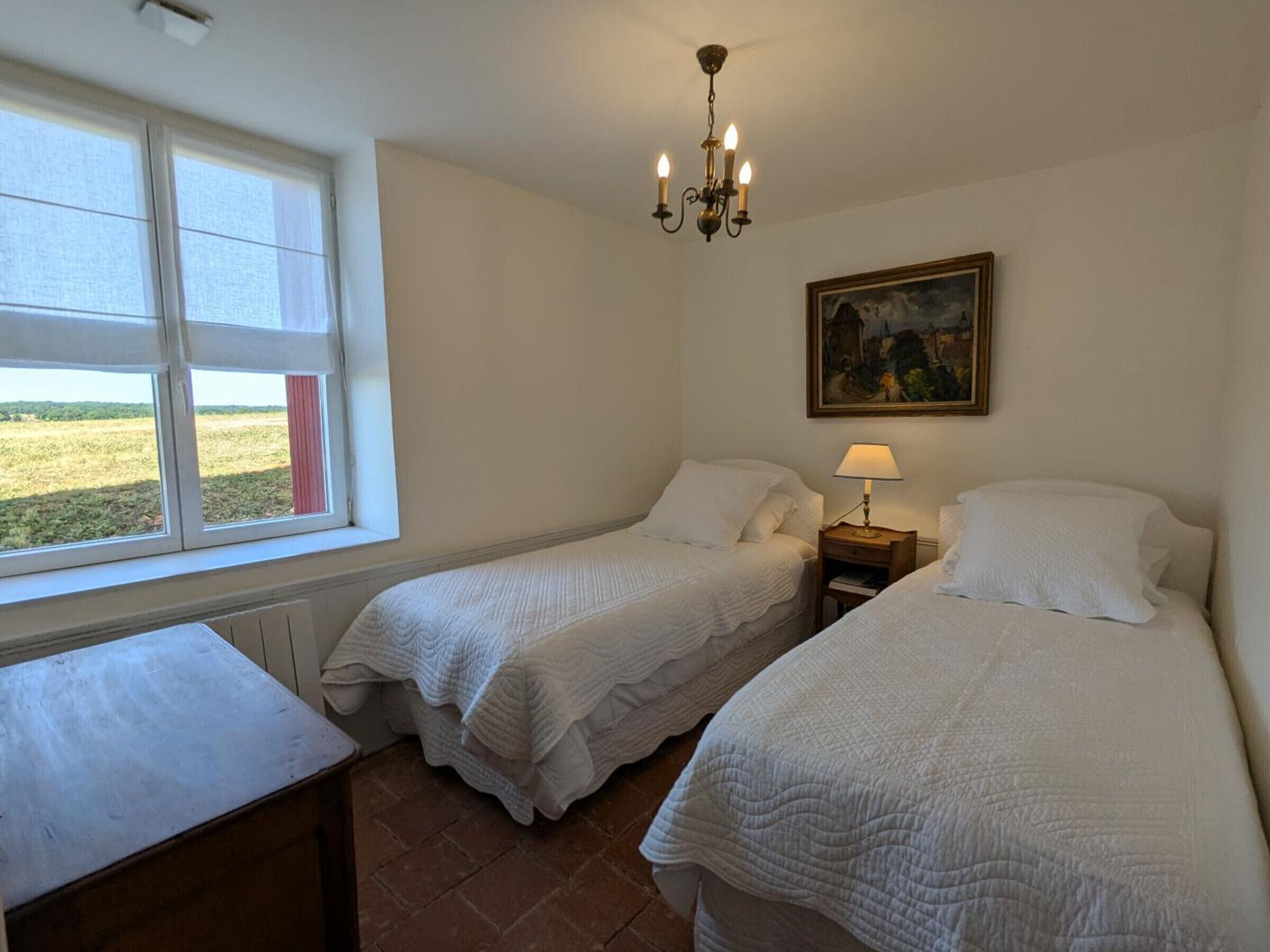 A bedroom with two single beds, a nightstand with a lamp, a painting on the wall, and a window with a view of the countryside.