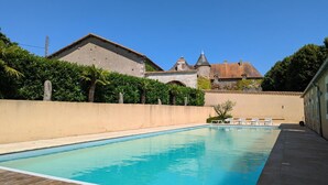 Ein Schwimmbecken mit Holzterrasse, umgeben von einer niedrigen Mauer und einem Heckenstreifen. Im Hintergrund ein Gebäude mit Natursteinfassade und einem Turm.