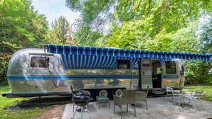 Una caravana antigua con toldo azul, una parrilla para barbacoa y mobiliario exterior.