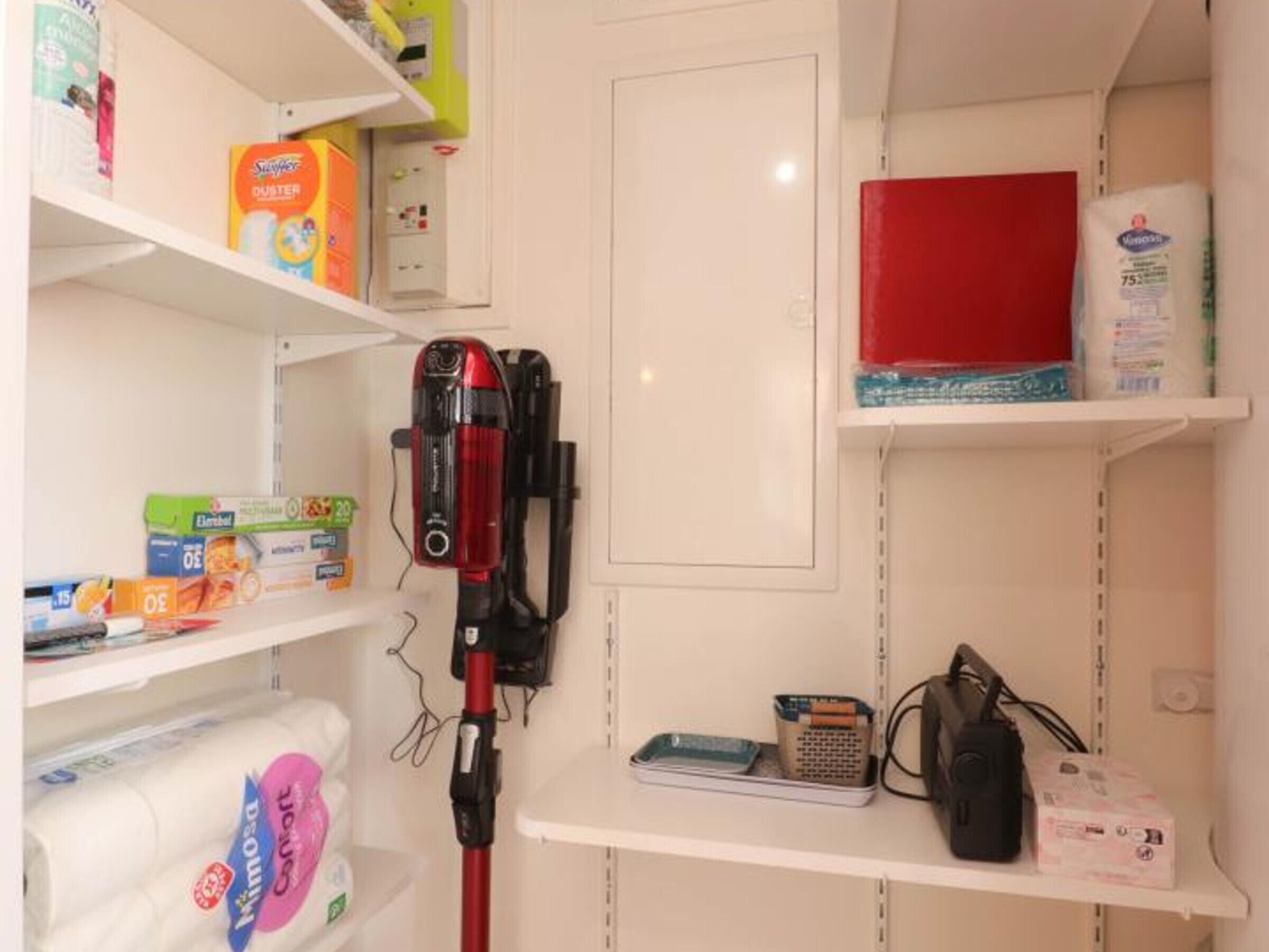 A storage area with shelves containing various household items like cleaning supplies, a red box, and a black bag.
