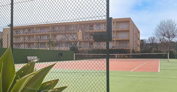 Una cancha de tenis con una red, rodeada por una cerca de malla metálica. Al fondo, se observa un edificio de varios pisos con balcones.
