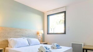A modern bedroom with a wooden headboard, a large bed with white linens, two bedside tables with lamps, and a window providing natural light. - Charming apartment in La Favière, pets allowed, near beaches (Bormes-les-Mimosas)