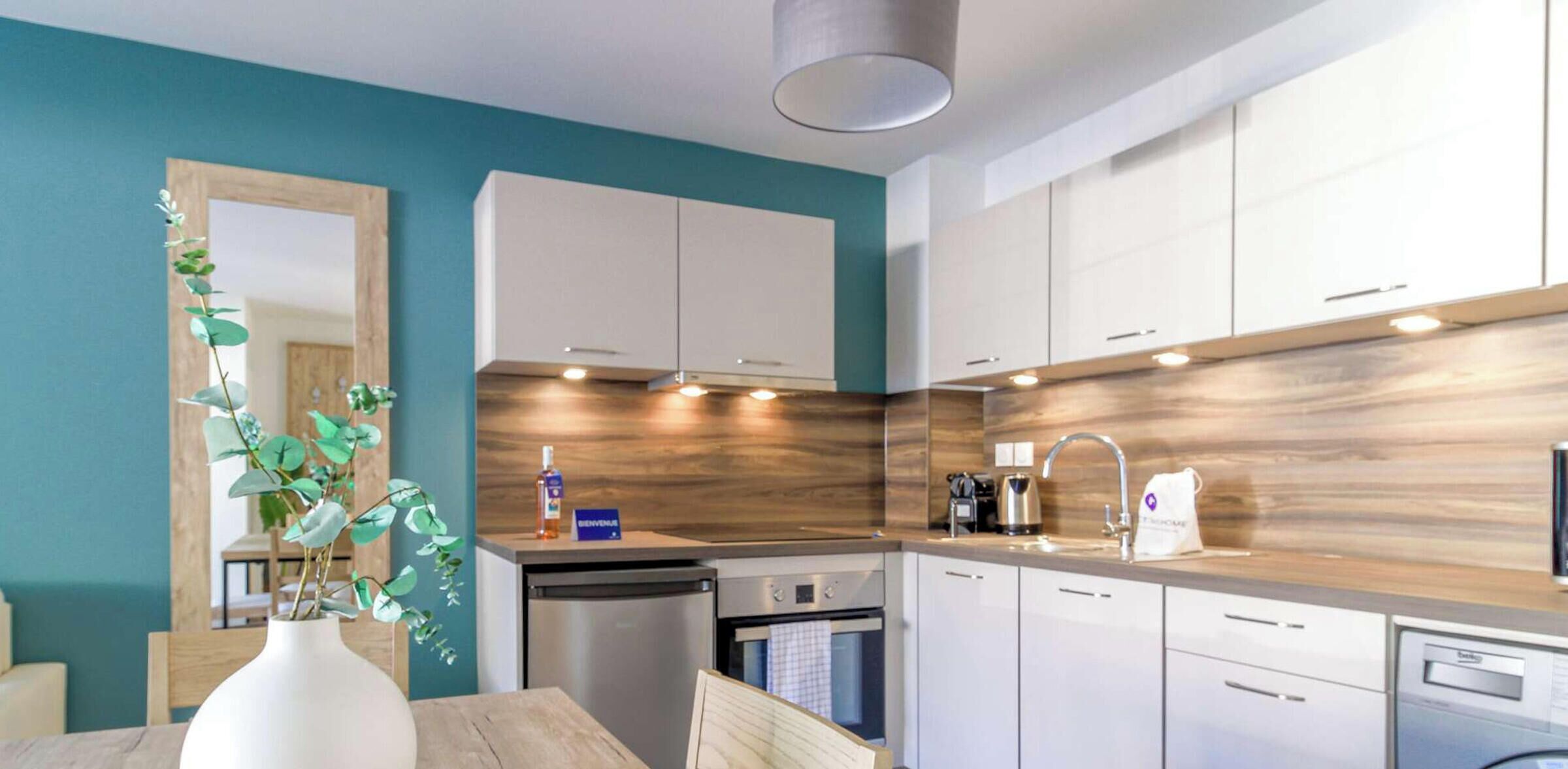 Een moderne keuken met witte kastjes, een houten spatwand, en een eethoek met een vaas groene bladeren.