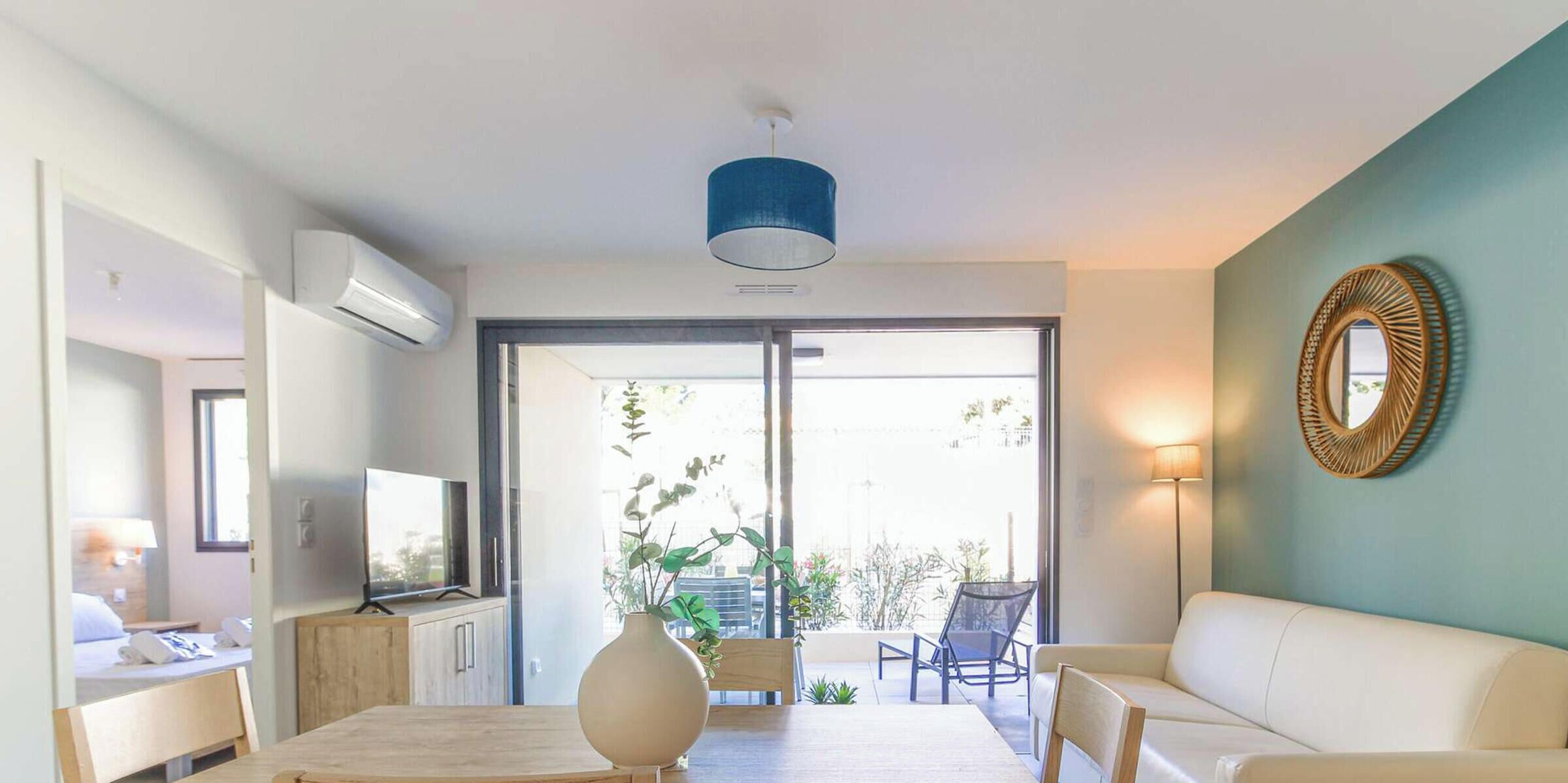 A modern living room with a dining table, chairs, a sofa, and a TV. There is a large window with a view of the outdoors.
